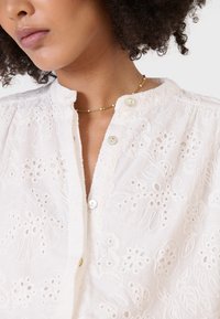 Blusa bianca ricamata con motivi floreali, colletto rotondo, chiusura con bottoni e tessuto testurizzato con dettagli in occhio di pernice.
