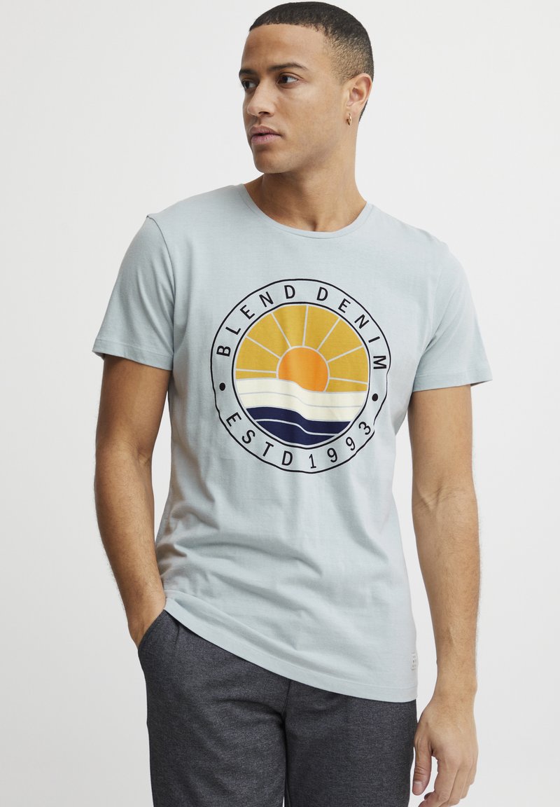 Lyseblå kortermet t-skjorte med et rundt motiv som viser en gul sol, bølger i hvitt og marineblått, og teksten "BLEND DENIM ESTD 1993".