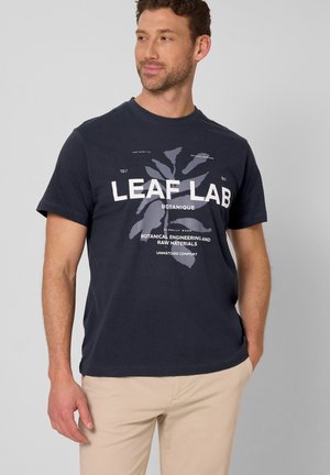 Uomo che indossa una maglietta grafica blu navy con la scritta "LEAF LAB" e pantaloni beige, in piedi con una mano in tasca, che guarda di lato su uno sfondo neutro.