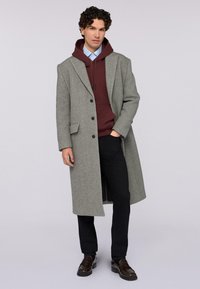 Grauer langer Mantel aus strukturiertem Wolle, mit Reverskragen, schwarzen Knöpfen und getragen über einem kastanienroten Hoodie und schwarzen Hosen.