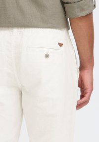 White linen pants featuring a back pocket with a button closure and a small brown logo patch above the pocket.
