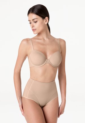 Donna che indossa un reggiseno beige modellato e mutandine a vita alta senza cuciture, con i capelli raccolti, in piedi davanti a uno sfondo bianco uniforme.