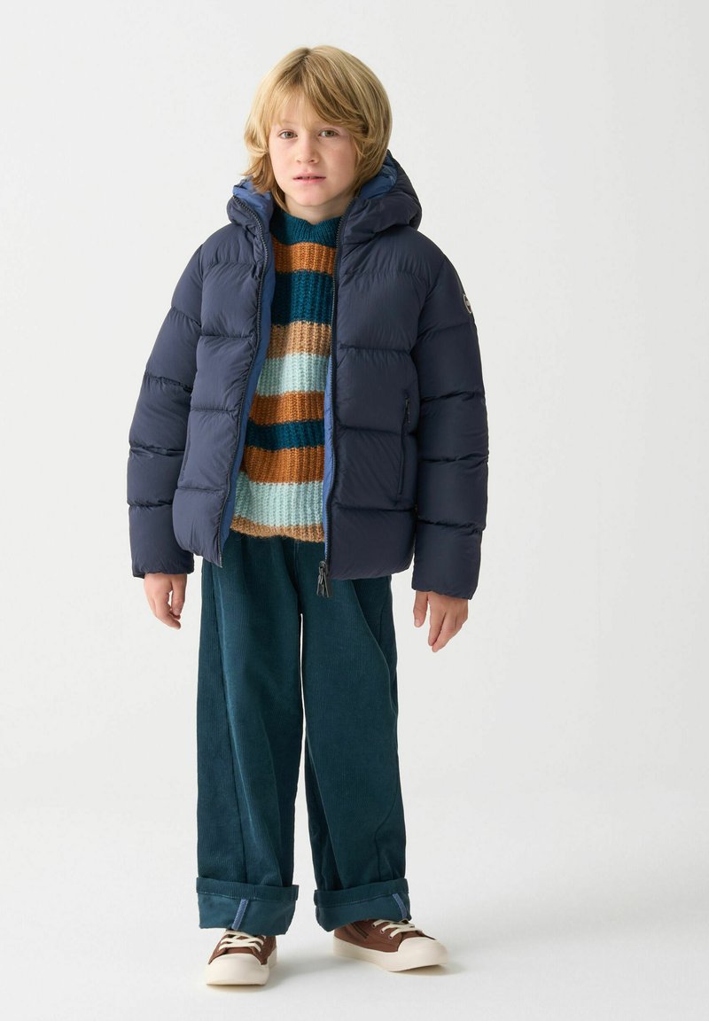 Giubbotto imbottito blu navy con cappucci, caratterizzato da sezioni trapuntate. Indossato sopra un maglione a righe in maglia e abbinato a pantaloni in velluto a coste teal. Sneakers casual.