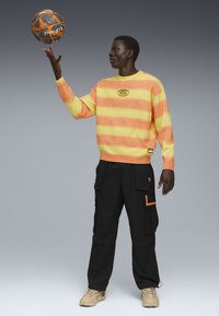 Man in een oranje en gele gestreepte trui draait een zwarte en oranje basketbal op zijn vinger, gekleed in zwarte cargobroeken en beige sneakers.