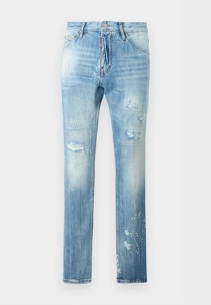 Young man with curly hair wearing a light blue short-sleeve polo shirt, distressed light blue jeans, and white sneakers, standing with one hand in pocket.