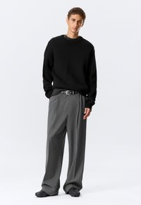 Jeune homme debout les mains dans les poches, portant un pull noir, un pantalon large gris à fines rayures, une ceinture et des chaussures noires texturées sur fond blanc.