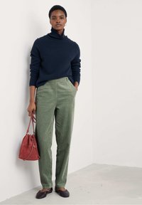 Ženska oblečena v mornarsko modro pulover s turtleneck izrisom in zelene žametne hlače, drži rdečo semiš torbico v obliki vedra, stoji ob beli steni.