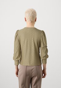 Olive green blouse with puffed sleeves, featuring a textured design of diamond shapes and perforations, paired with beige trousers.