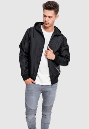 Sort letvægtsjakke med hætte og frontlynlås, ribbede manchetter, båret over en hvid t-shirt og parret med lysegrå jeans med strukturerede paneler.