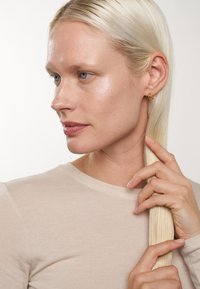 Guldiga hoops, slät hy, långt rakt hår, iklädd en beige långärmad topp. Neutral bakgrund framhäver ansiktsdragen.