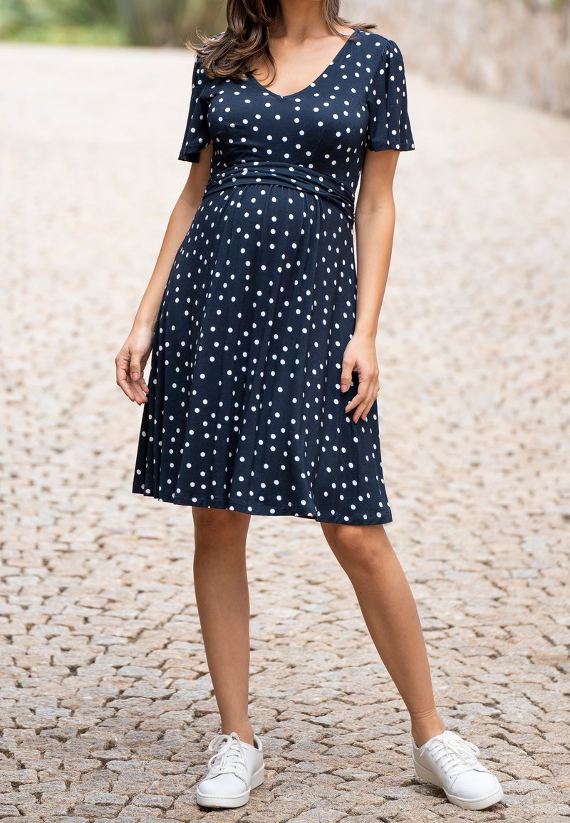 Femme portant une robe noire mi-longue à pois blancs et des baskets blanches, debout sur un chemin pavé en plein air.