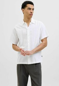 Camicia bianca a maniche corte con motivo testurizzato, colletto e taglio dritto, abbinata a pantaloni scuri sartoriali.
