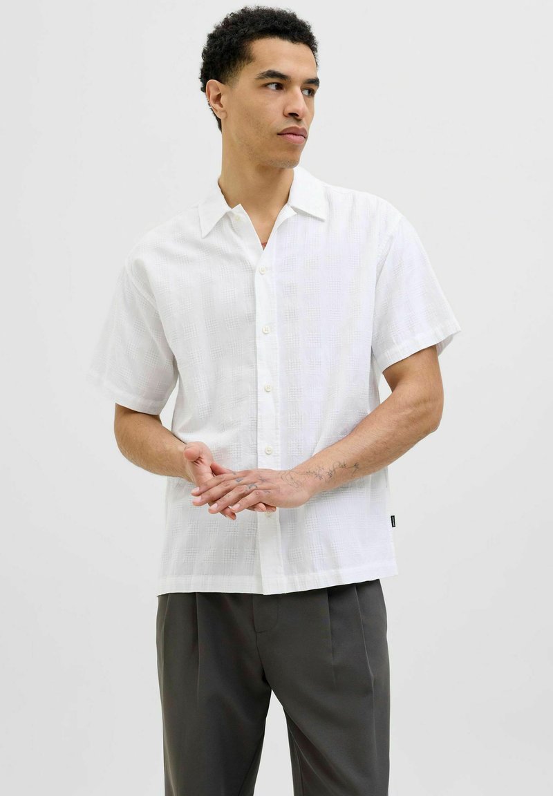 Camicia bianca a maniche corte con motivo testurizzato, colletto e taglio dritto, abbinata a pantaloni scuri sartoriali.