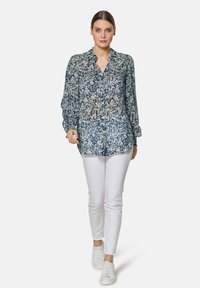 Bluse mit floralem Muster in Blau und Creme, mit durchgehender Knopfleiste, langen Ärmeln und lockerer Passform, kombiniert mit weißen Slim-Pants und weißen Sneakers.