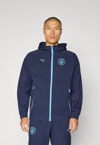 Donkerblauwe hoodie met een lichtblauwe rits en accenten. Voorzien van een Manchester City-embleem op de borst en elastische manchetten voor een goede pasvorm.