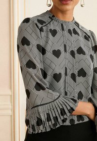 Blusa in bianco e nero con un motivo a cuori geometrici, caratterizzata da una rouches plissettata sulle maniche e un colletto rotondo.