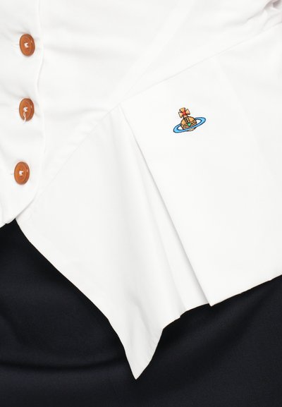 Hvid bluse med spids kant, prydet med brune knapper og et farverigt broderet design af en krone ovenpå en planet, sat mod en mørk baggrund.