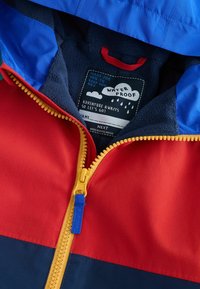 Veste imperméable à blocs de couleur avec des panneaux rouges, bleus et marins. Dotée d'une fermeture éclair jaune et d'une doublure en polaire. Étiquette d'entretien visible à l'intérieur.