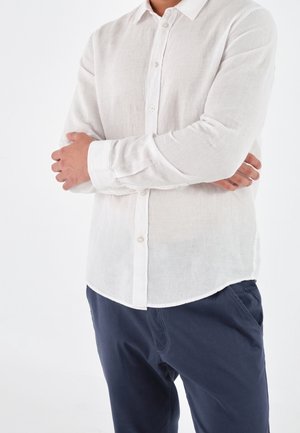 Personne portant une chemise blanche à boutons avec manches longues et un pantalon bleu marine, bras croisés sur la poitrine devant un fond clair uni.