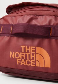 Sac de voyage en tissu bordeaux avec le logo orange "The North Face". Dispose d'une fermeture éclair sur le dessus et de bretelles texturées pour un transport pratique.