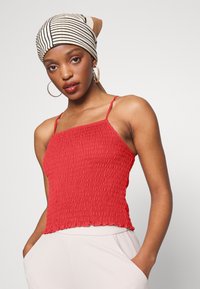 Camisola smock vermelha com alças finas, apresentando uma superfície texturizada e uma bainha em forma de ondas. Combinada com um lenço de cabeça riscado e brincos de argola.
