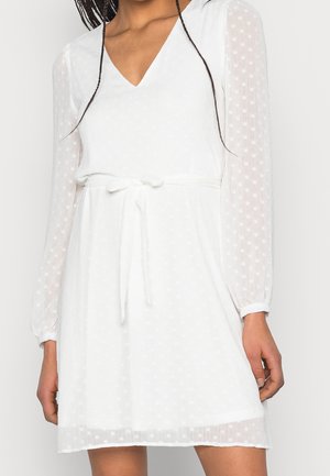 Femme portant une robe blanche à col en V avec des manches longues transparentes, des pois texturés et une ceinture nouée à la taille.