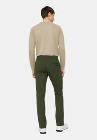 Camicia beige a maniche lunghe e pantaloni verdi con cintura elastica, caratterizzati da una tasca posteriore con logo. Le scarpe sono bianche con suole nere.