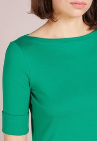 Grön, figurnära topp i slätt tyg. Har en bred halsringning och trekvartsärmar med uppvikta manschetter. Minimala sömmar synliga.