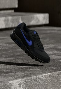 Zwarte Nike Air Max sneaker met een blauwe swoosh, gestructureerd bovenwerk, gedempte zool en zichtbare luchtunit, geplaatst op een grijs oppervlak.