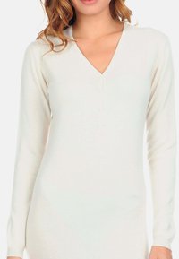 Haut en tricot blanc à manches longues avec un col en V et une patte de boutonnage, présentant une texture douce et une coupe ajustée. Aucun motif ni accent présent.