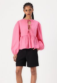 Top rosa a maniche lunghe con vita arricciata, apertura con fiocco nella parte anteriore e maniche a palloncino, abbinato a pantaloni corti neri con risvolti.