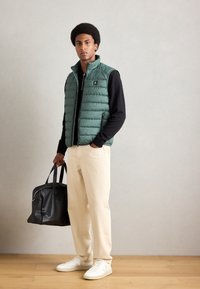 Grüne, puffy Weste mit hohem Kragen, Reißverschluss vorne und seitlichen Taschen, kombiniert mit einem schwarzen Langarmshirt, beige Hosen und weißen Sneakers.