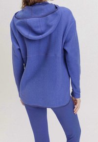 Lila fleecehoodie med en mjuk textur och en rymlig huva, med kontrasterande sömsömmar. Bärs med matchande lila leggings.
