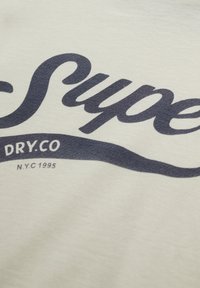 Tissu de couleur crème avec texte en écriture cursive bleu foncé "Super" et texte plus petit "DRY.CO N.Y.C 1995" en dessous. Texture légère, style décontracté.