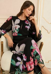 Čierna kvetinová šaty s dlhými, priehľadnými rukávmi. Obsahujú veľké ružové a fialové kvetinové vzory, výstrih v tvare kľúčovej dierky a splývavú siluetu.
