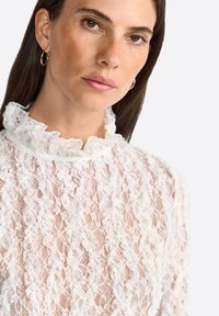 Primo piano di una donna con lunghi capelli scuri, che indossa un top di pizzo bianco con collo arricciato, mostrando il suo trucco naturale e un'espressione neutra.