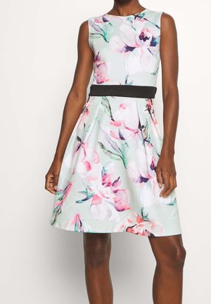 Sukienka bez rękawów, do kolan, w pastelowy kwiatowy wzór z czarnym paskiem w talii, noszona przez ciemnoskórą osobę stojącą na jednolitym tle.