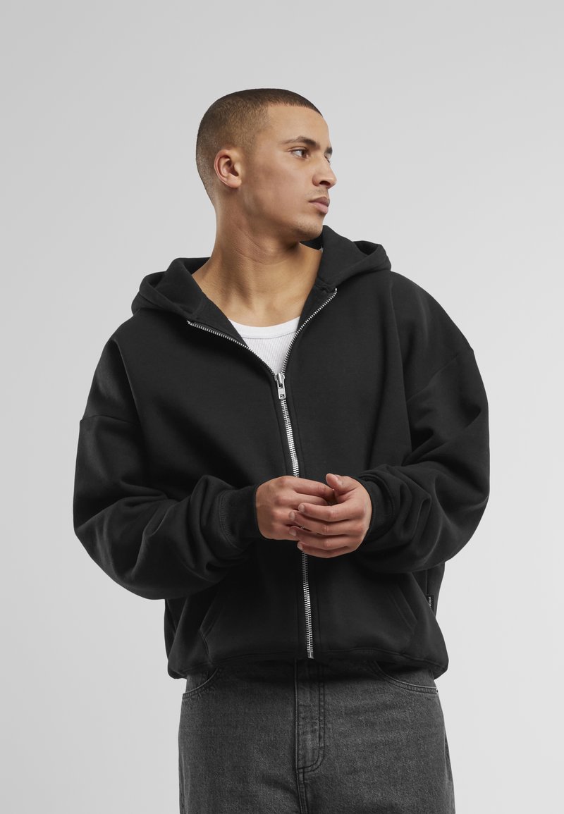 Schwarze Zip-Up-Hoodie mit einer geräumigen Kapuze, langärmligen Bündchen und Fronttaschen, getragen über einem weißen Tanktop aus strukturiertem Stoff.