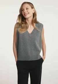 Szara bezrękawnikowa bluzka dzianinowa z dekoltem w kształcie litery V, prążkowanym dołem i miękką teksturą. Nosi się ją na czarnych spodniach, charakteryzujących się minimalistycznym wzornictwem i stonowaną kolorystyką.