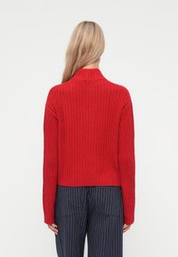 Rød ribbstrikket genser med høy krage og lange ermer, med et kort design. Brukt sammen med navy stripete bukser.
