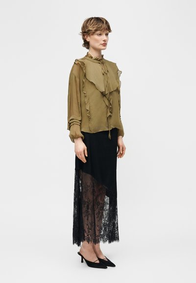 Γυναίκα που φοράει πουκάμισο ελιάς με βολάν και μαύρη ασύμμετρη φούστα με διαφανές δαντελένιο τελείωμα, συνδυασμένα με μαύρα παπούτσια με χαμηλό τακουνάκι.