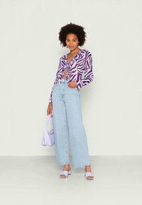 Blusa padrão roxa e branca com laço na frente, combinada com jeans de perna larga azul claro e acessórios lilás, incluindo uma mala e sandálias.