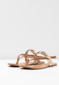 Flade sandaler med et fletværk i beige, brune remme og en glat tan-sål. Enkel design med minimal udsmykning.
