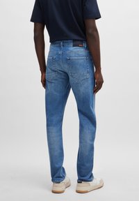 Man som bär ljusblå denimjeans, svart skjorta och vita sneakers, står vänd bort från kameran mot en enkel grå bakgrund.