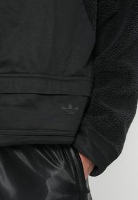 Črn pulover z flis teksturo na eni strani in gladkim materialom na dnu. Ima toniran Adidas logo na robu.