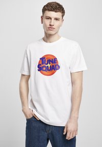 Hvit bomullst-skjorte med et rundt grafisk motiv i oransje og blått med teksten "TUNE SQUAD" i fet skrift. Korte ermer og en avslappet passform.
