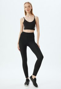 Zwarte atletische crop top met dunne bandjes en witte accenten aan de zijkanten, gecombineerd met hoge zwarte leggings. Het model draagt zwarte sneakers.