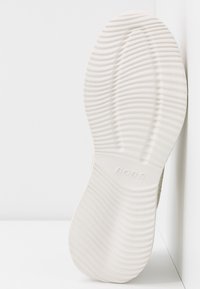 Suola di scarpe bianca con motivo di grip ondulato testurizzato e il nome del marchio "BOBS" impresso al centro, in posizione verticale contro un angolo bianco.