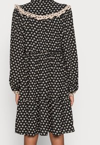 Robe noire à motif floral blanc, col à volants, manches longues, taille cintrée et jupe à volants. Tissu doux avec un design fluide.