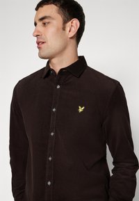 Camisa de pana marrón de manga larga con cuello abotonado. Presenta un pequeño logotipo amarillo bordado en el pecho. Botones de color gris claro.